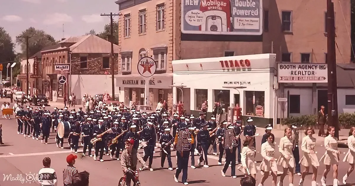 A marching band in blue uniforms parades down a small-town street with vintage storefronts and a Texaco gas station.