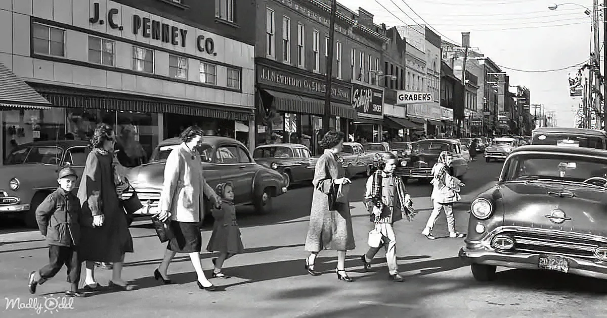 Pedestrians cross a busy street in front of J.C. Penney Co., surrounded by classic cars and storefronts.