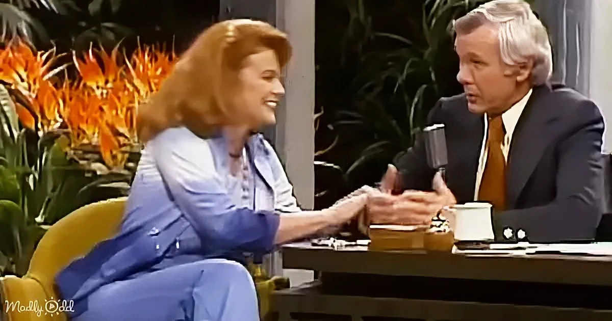 A woman in a blue suit is seated, smiling, and holding hands with a man in a suit during a talk show.