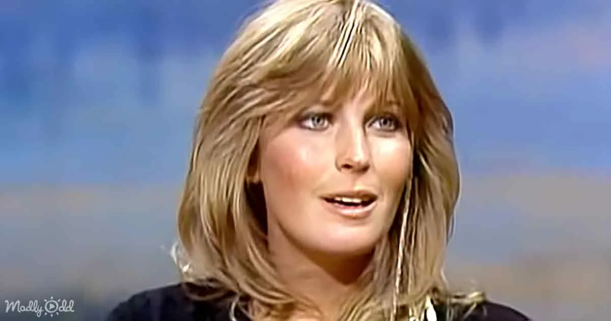 A person with blonde hair and bangs, wearing a dark top, is speaking against a blurred background.