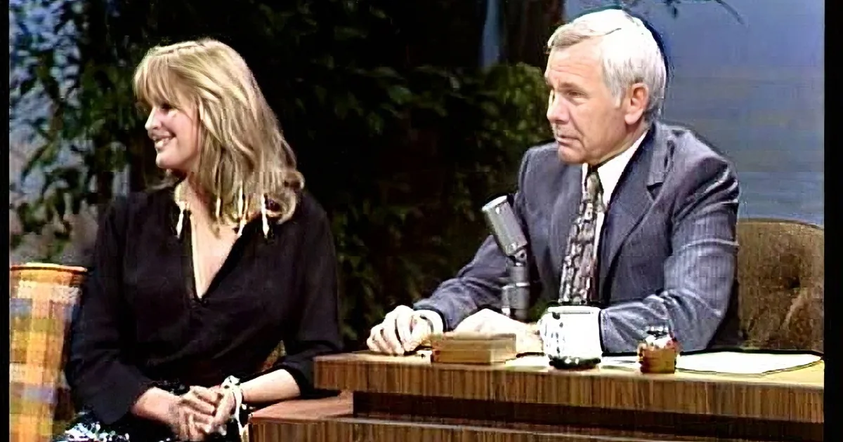 A woman with blonde hair smiles while seated at a table with a man in a suit, both engaged in conversation.