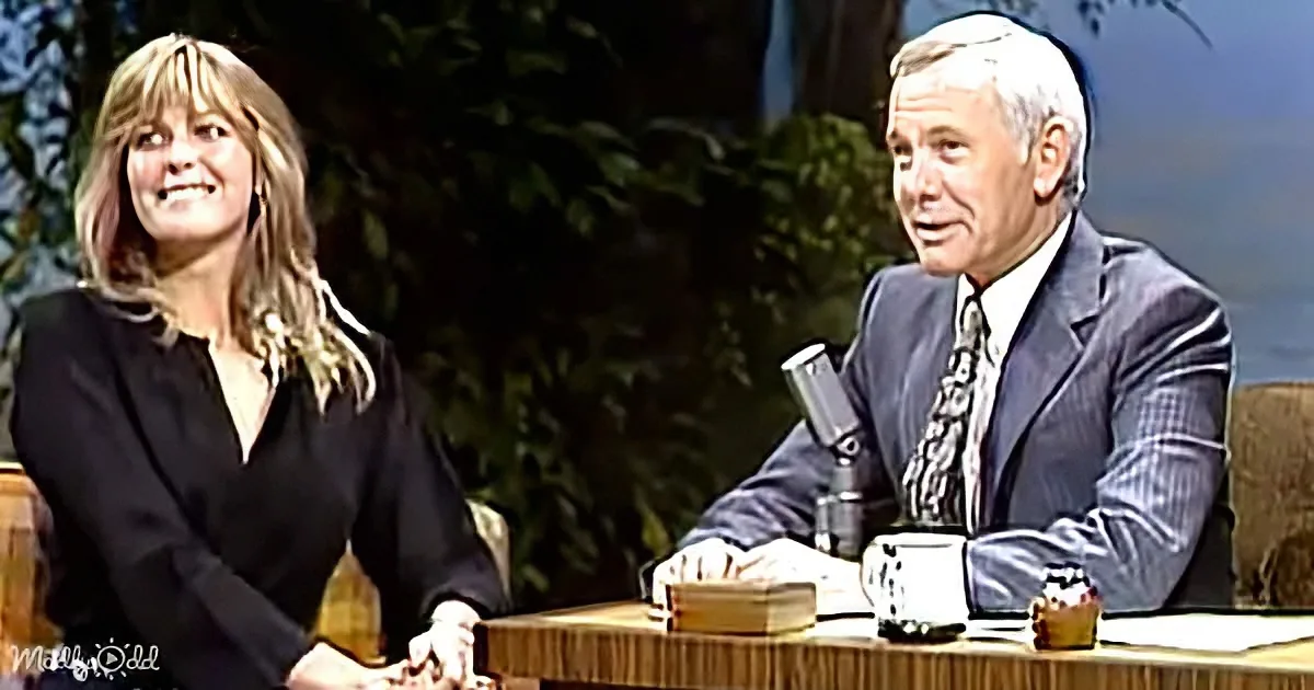 A woman smiles while seated at a table with a man in a suit, both appearing to be in a talk show setting.