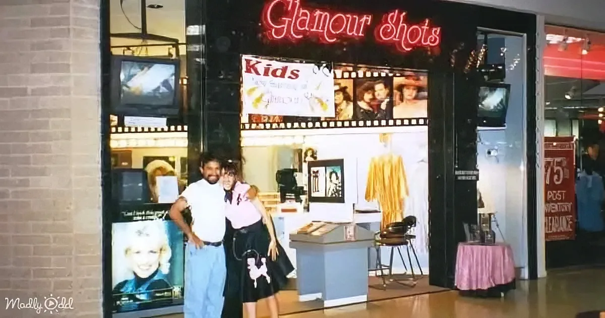 Two people pose in front of a "Glamour Shots" photo booth with a neon sign and vintage decor.