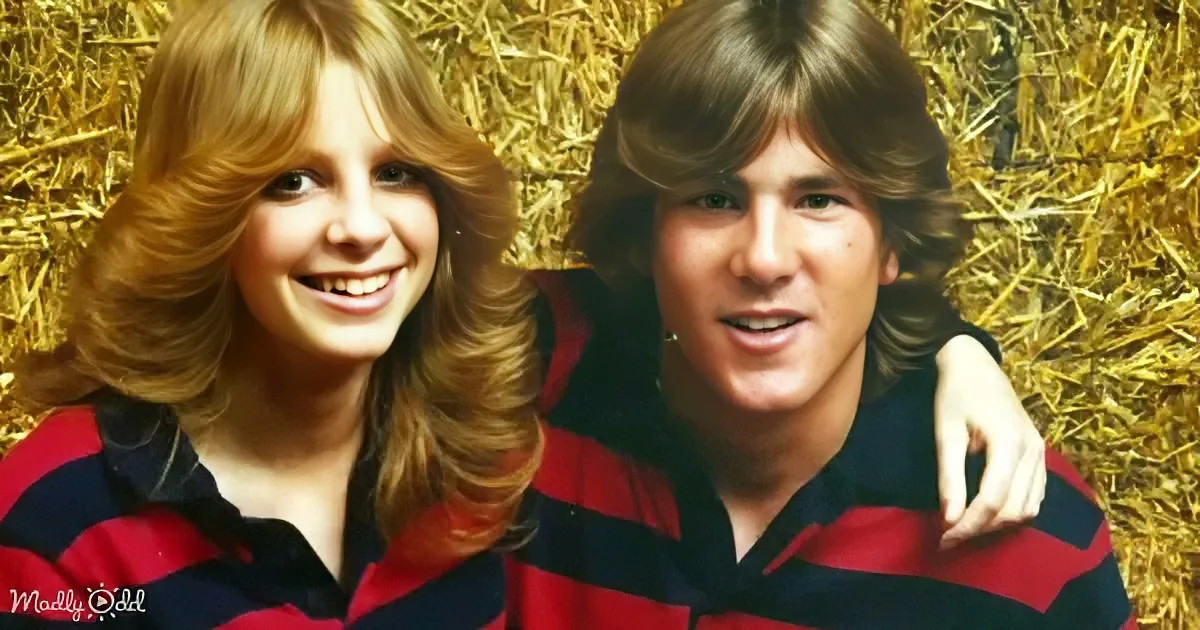 A smiling couple in matching red and black striped shirts stands against a hay background.