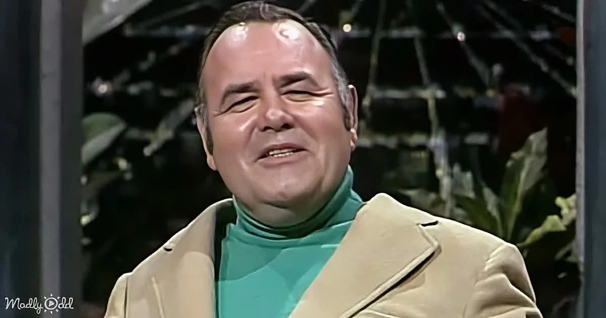 A man in a beige coat and teal turtleneck smiles with his eyes closed, surrounded by a dark, plant-filled background.