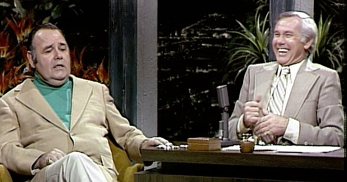 A man in a beige suit sits at a desk with a microphone, interviewing another man in a green shirt and beige coat.