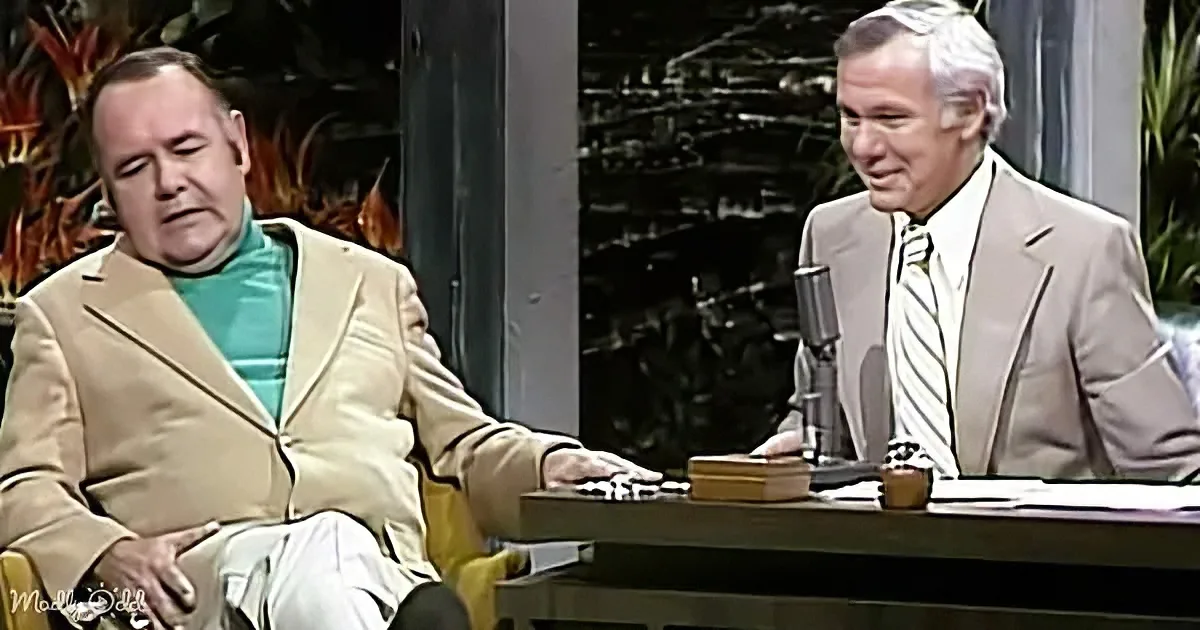 A man in a beige suit sits at a desk with a microphone, engaging in conversation with another man in a green shirt.