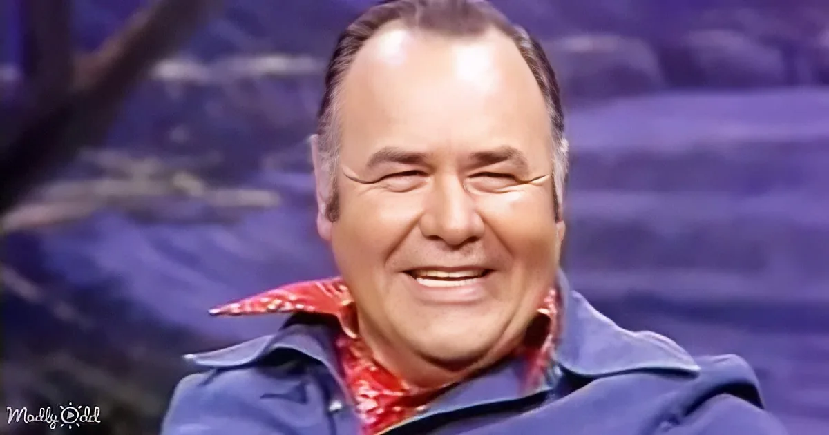 A man in a blue jacket and red shirt smiles widely, eyes closed, against a purple background.