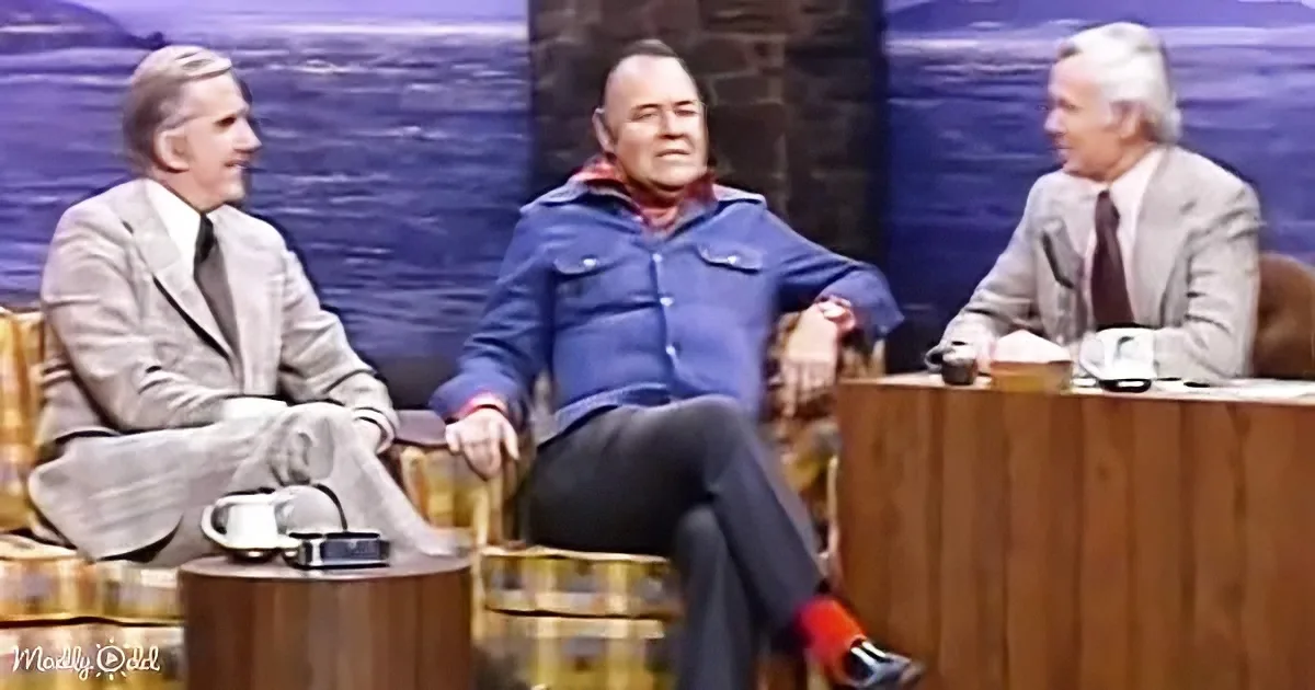 A man in a blue jacket sits relaxed, legs crossed, on a gold chair, engaged in conversation with two others on a talk...
