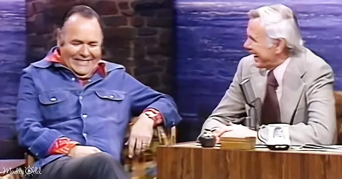 Two men are seated, one in a blue jacket smiling, the other in a suit appearing surprised or amused.