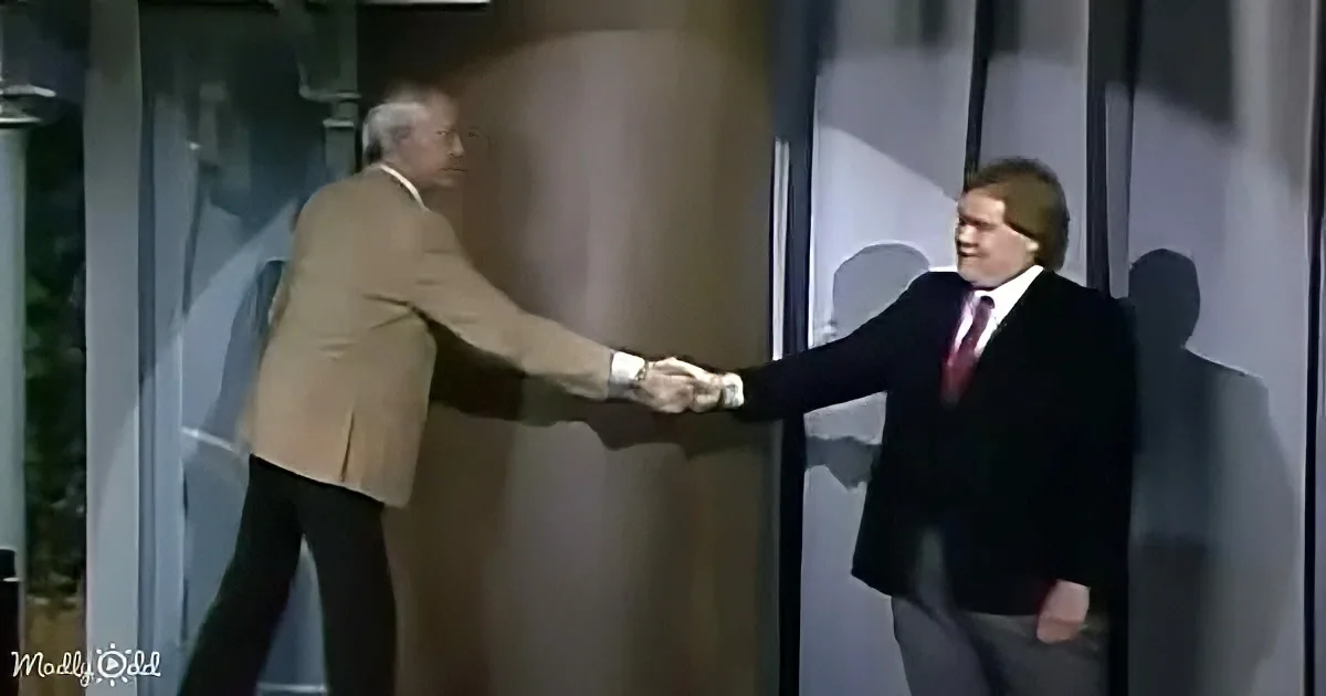 Two men in suits shake hands in a formal setting, one in a tan suit, the other in black.