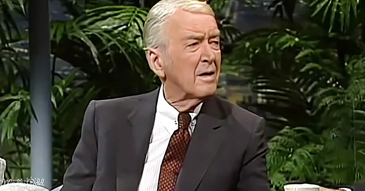 An older man in a suit and tie sits against a backdrop of greenery, appearing to speak.