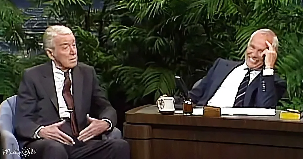 Two men in suits, one gesturing, seated on a talk show set with a desk and plants in the background.