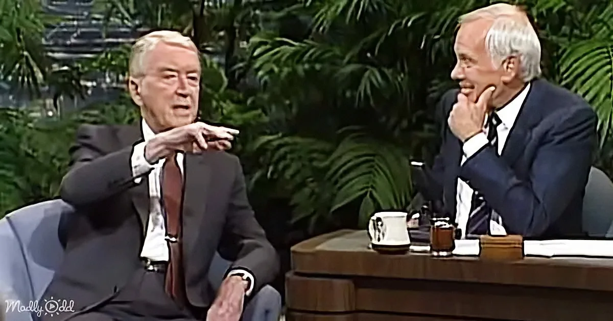 Two men in suits are seated, one gesturing while the other listens, with a coffee mug and papers on a desk.