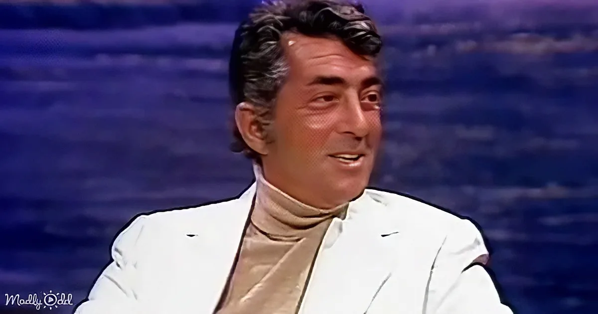 A man in a white suit and turtleneck smiles against a blue background.