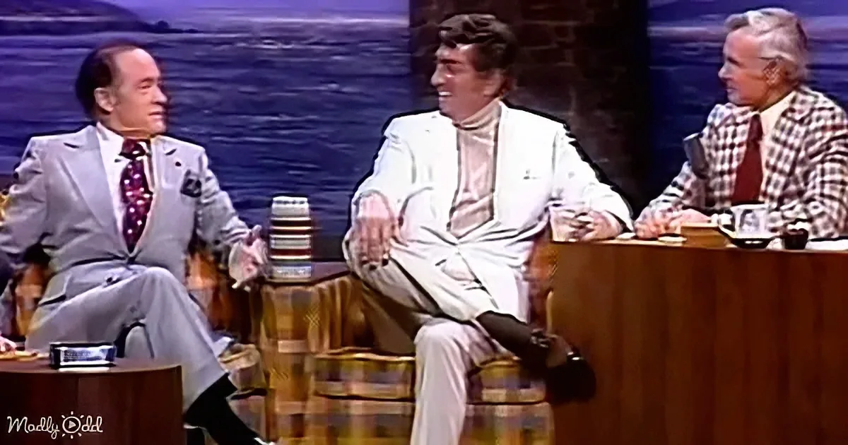 Three men in suits sit at a desk, one in a white suit appears relaxed, others engaged in conversation.
