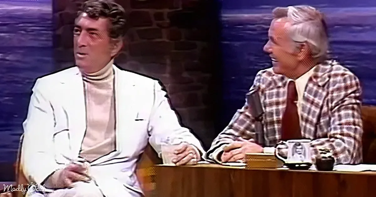 Two men sit at a table, one in a white suit, the other in a plaid shirt, engaged in conversation.