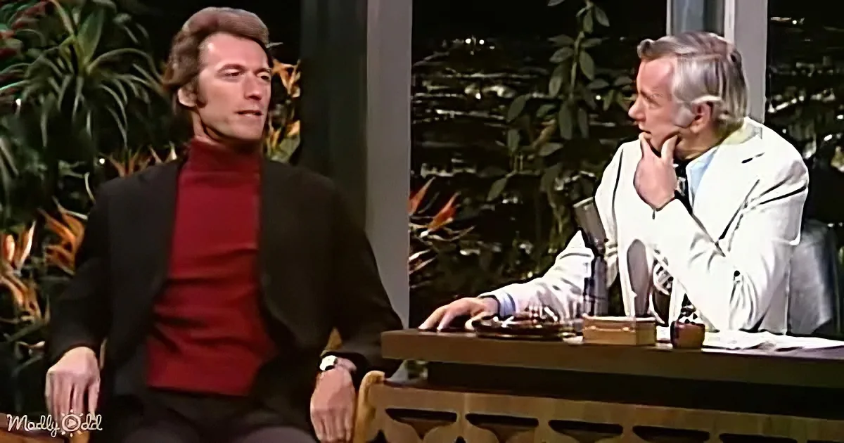 Clint Eastwood in a red turtleneck and black suit sits on a talk show set, facing a host in a white suit.