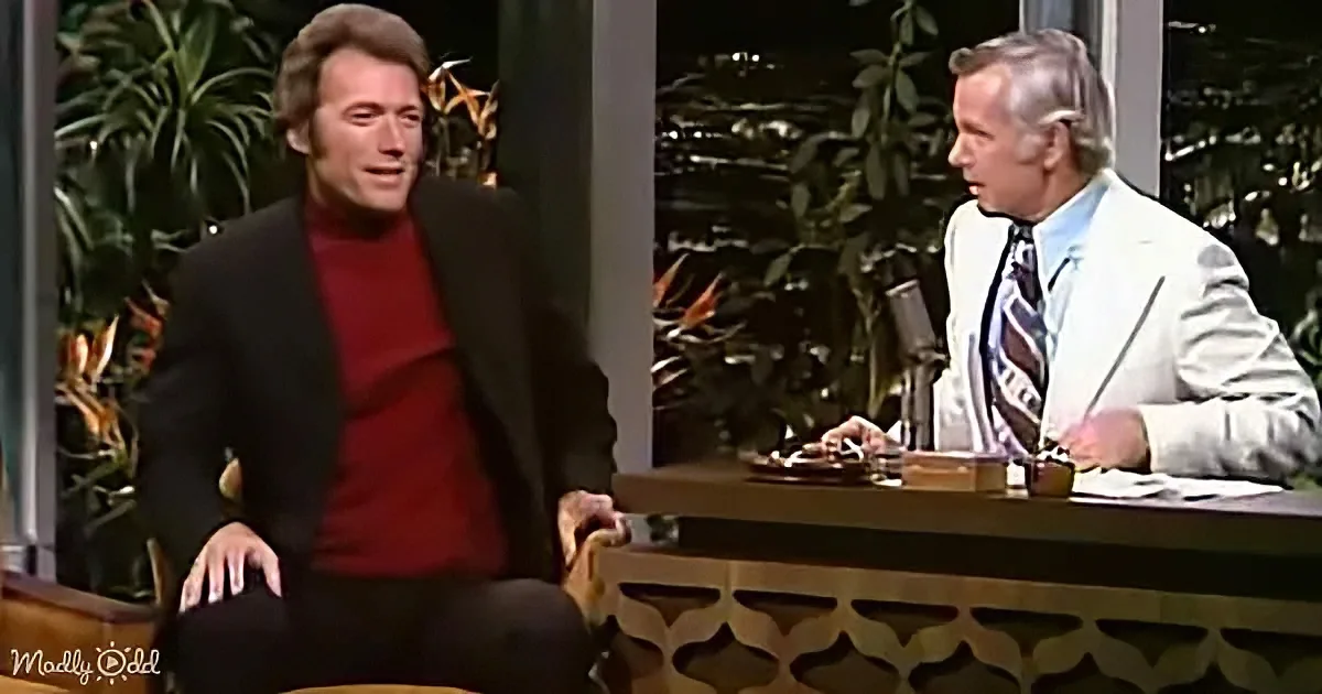 Clint Eastwood sits in a chair, wearing a red sweater and black jacket, on a talk show set.