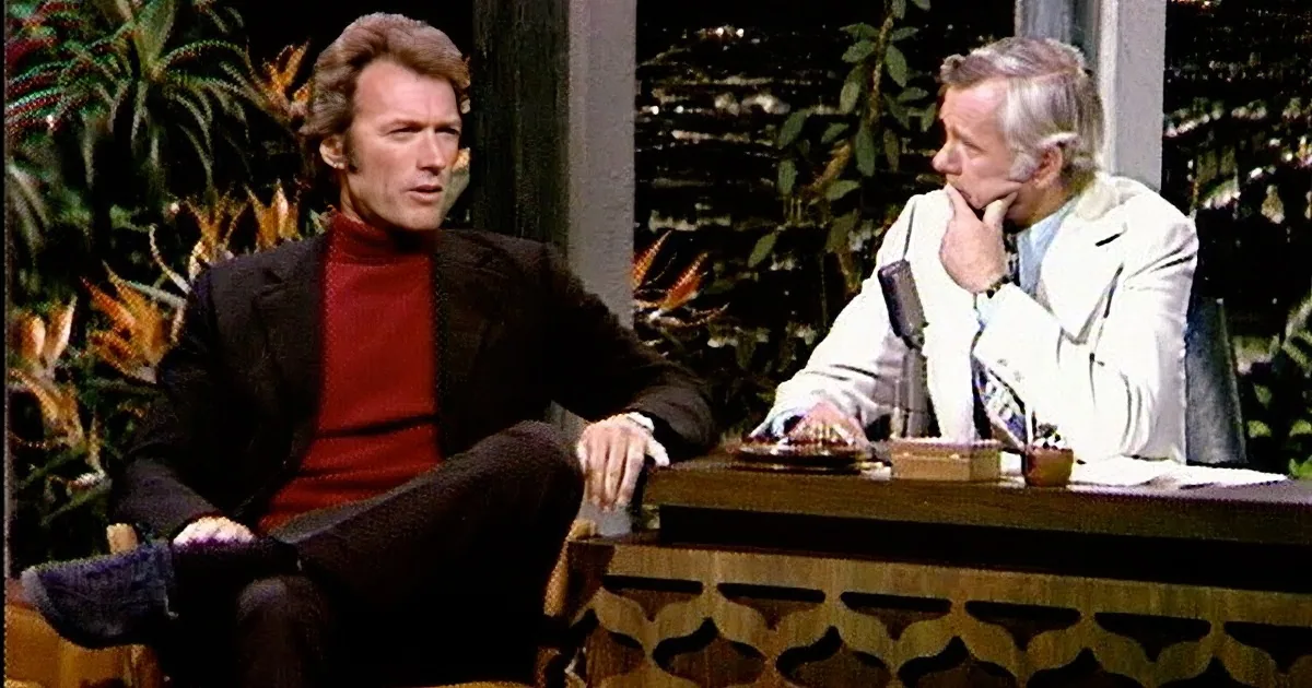 A man in a black suit sits with hands clasped, while another laughs in a white suit at a desk.