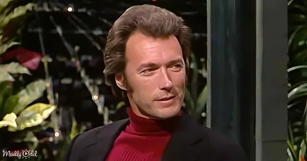 A man with short, wavy hair, wearing a red turtleneck and black blazer, stands in front of a backdrop with plants and lights.
