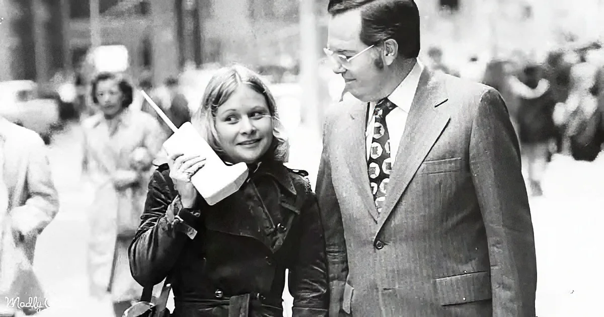 A woman holds a bulky cell phone while standing next to a man in a suit, both in a busy street scene.
