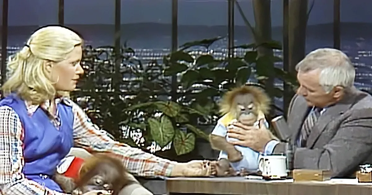 A man holds a baby orangutan while sitting with a woman on a talk show set.