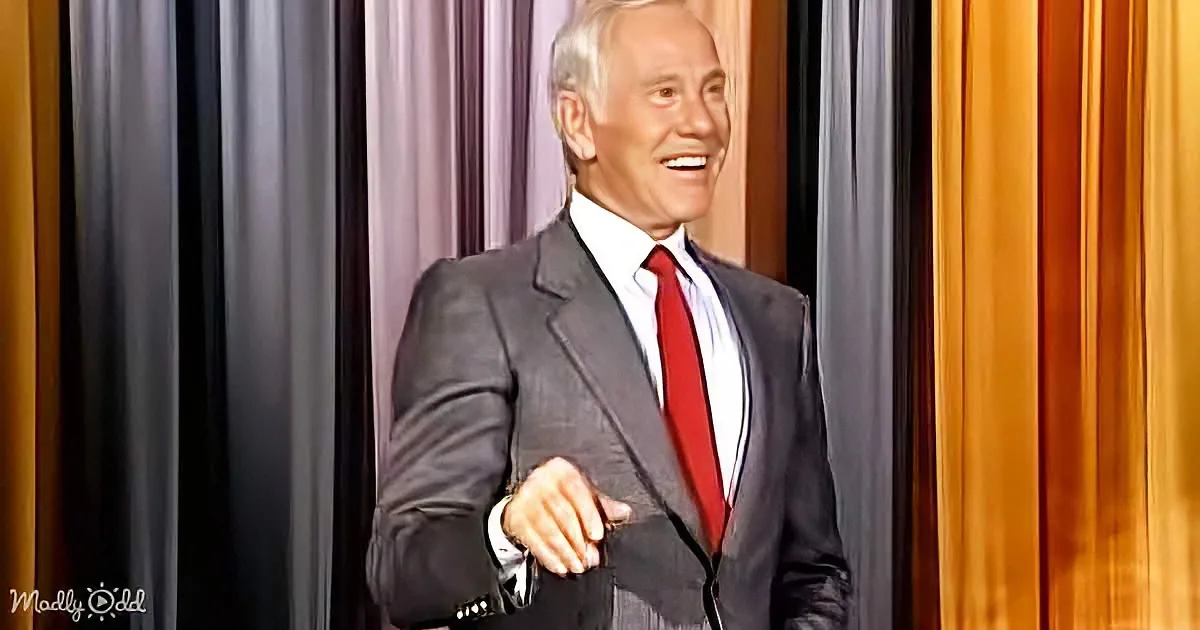 A man in a gray suit and red tie smiles, standing against a striped backdrop.