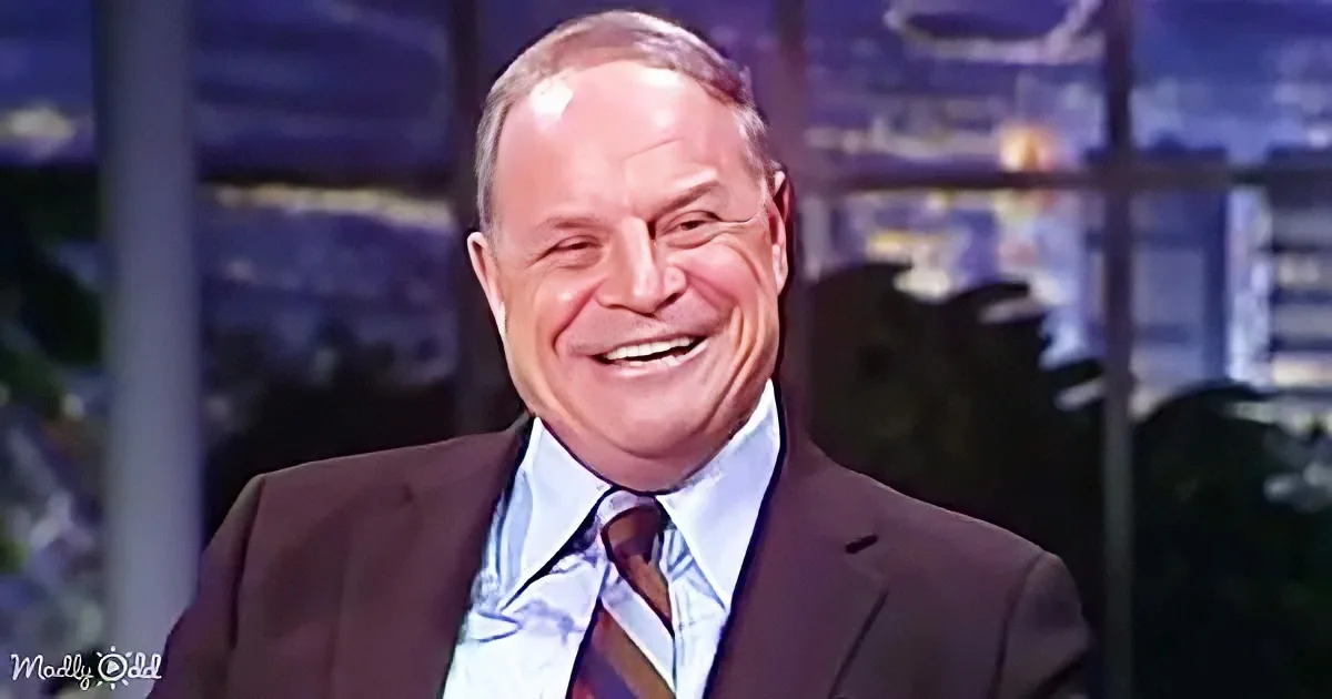 A man in a suit and tie smiles broadly, seated in a studio setting with blurred background.