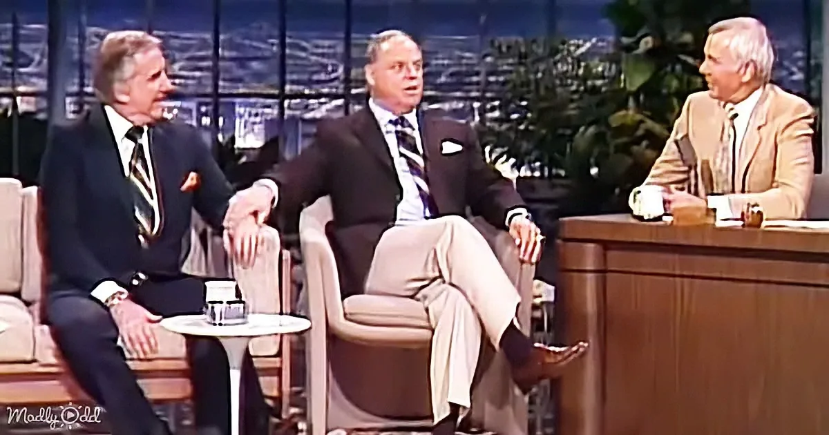 Three men in suits sit on a talk show set, engaged in conversation.