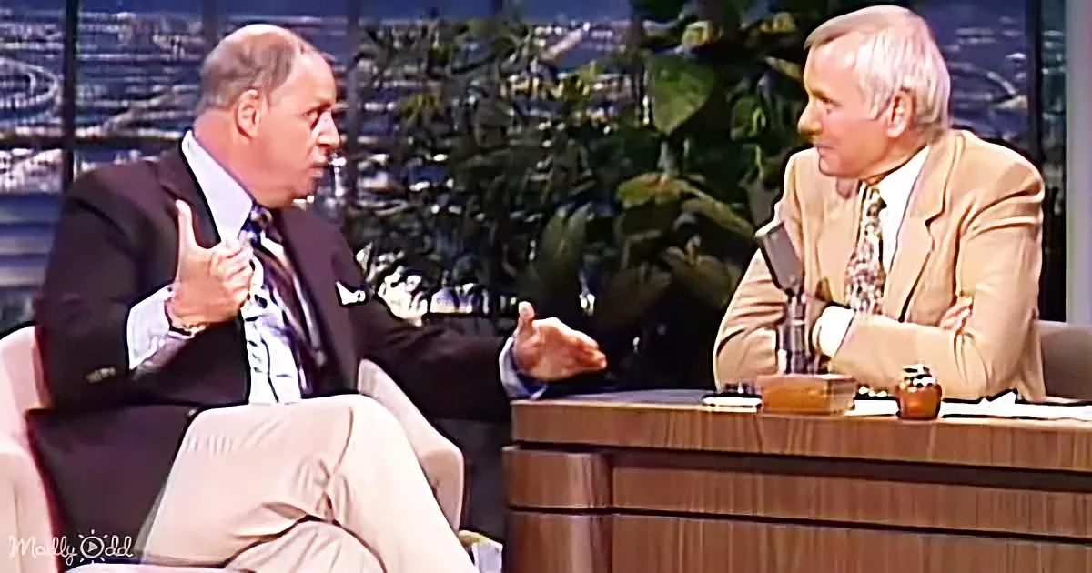 Two men in suits are seated, one gesturing with hands, in a talk show setting with a cityscape backdrop.