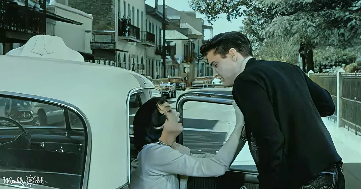 A man leans over a woman in a white dress, both near a vintage car, with a street scene in the background.
