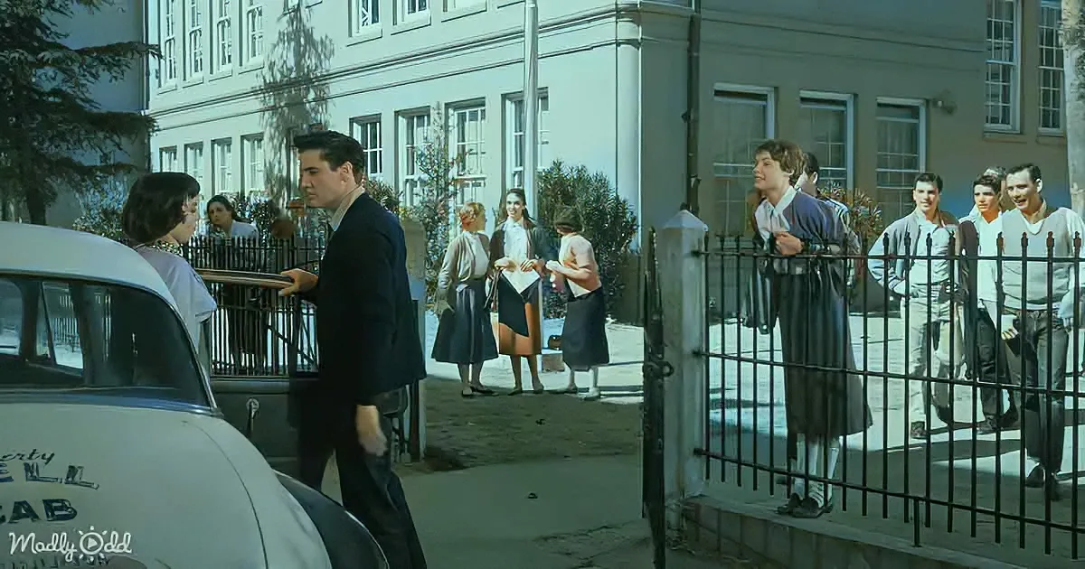 A man in a suit stands near a car, talking to a woman, while a group of people, including students, gather outside a school.