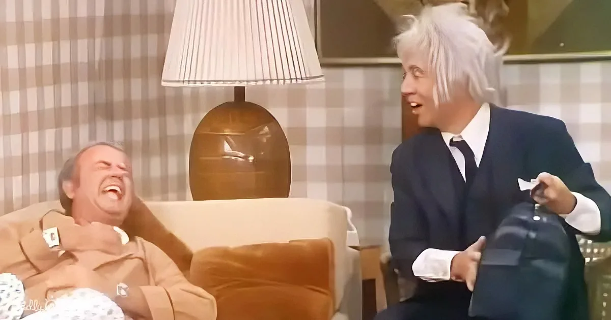 A man in a suit laughs while another in a brown coat sits on a chair, with a lamp in the background.