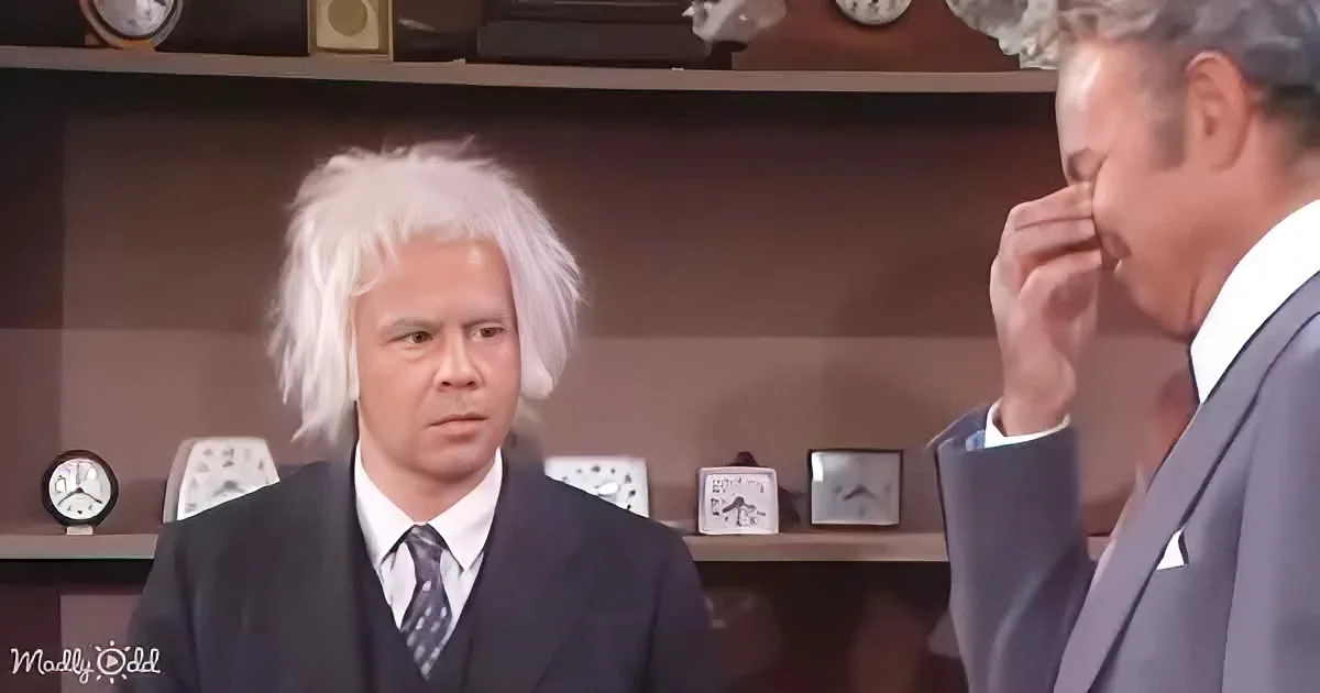 A man in a suit with white hair and a serious expression stands in front of a shelf with clocks.