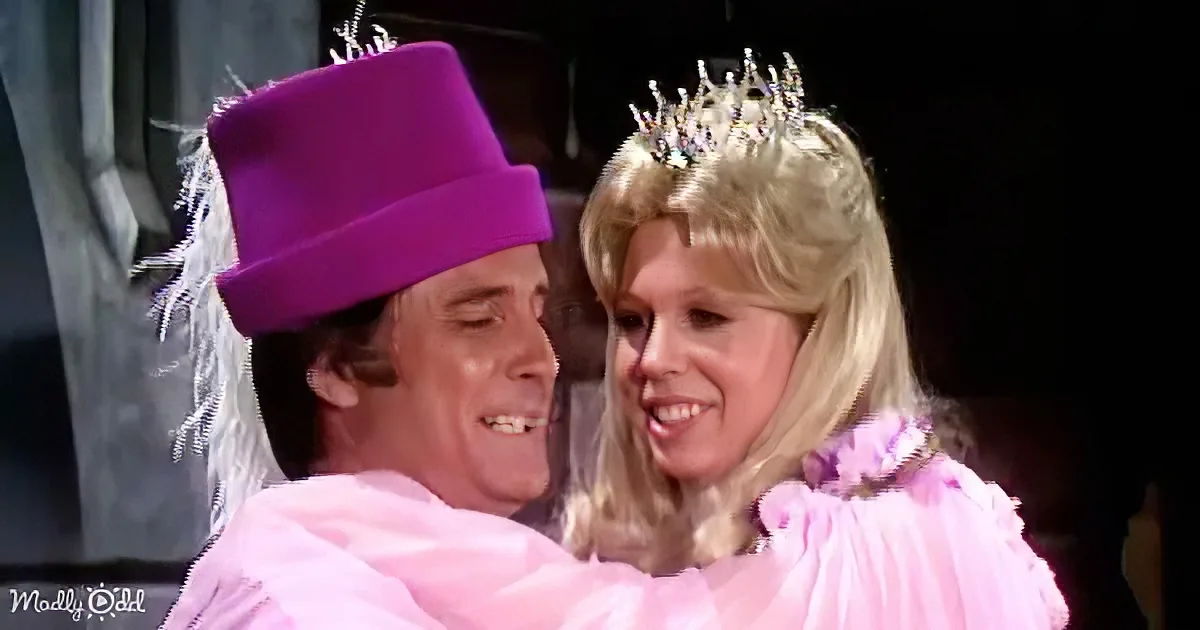 A man and woman in pink costumes, the man wearing a purple hat, the woman a tiara, share a joyful moment.