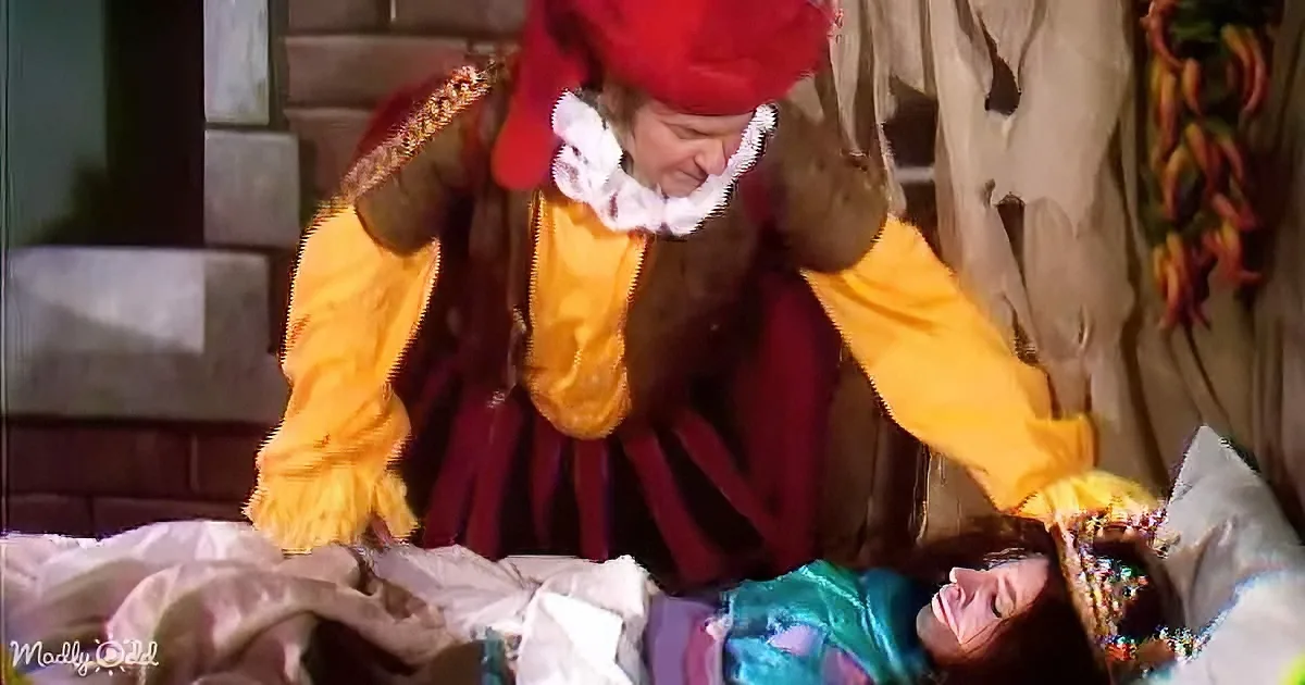 A person in a red and yellow costume leans over a table with a colorful cloth, possibly in a theatrical or festive setting.