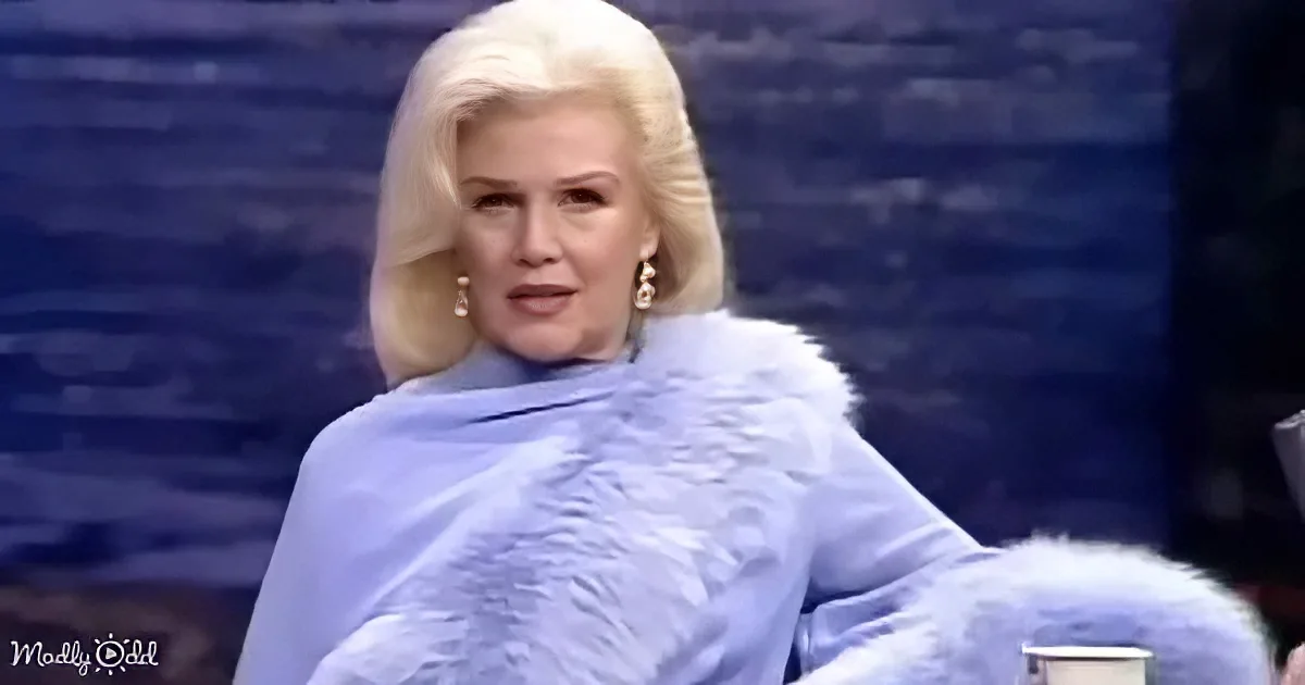 A woman with blonde hair and earrings, wearing a blue fur coat, is seated and appears to be speaking.