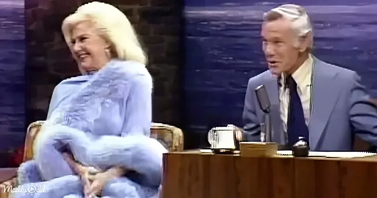 A woman in a blue fur coat sits on a chair, smiling, while a man in a suit sits at a desk with a microphone.