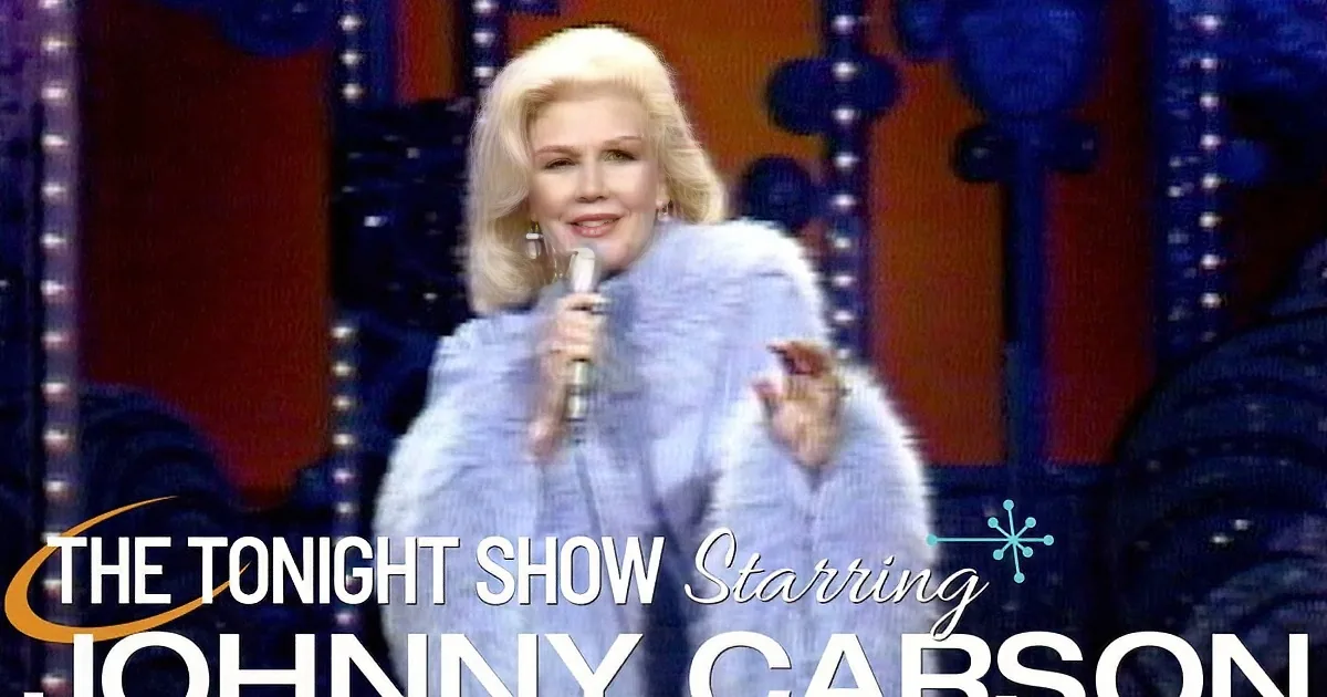 A woman in a light blue fur coat sings into a microphone on stage.