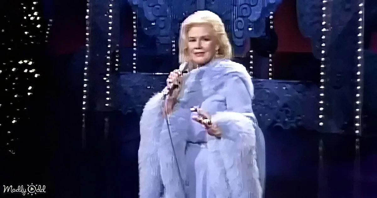 A woman in a white fur coat sings on stage, holding a microphone, with a dark, patterned background.