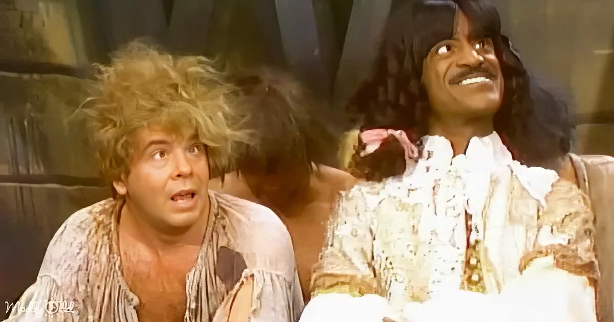 A man with wild, curly hair and a surprised expression is sitting next to another man in a white, patterned shirt.