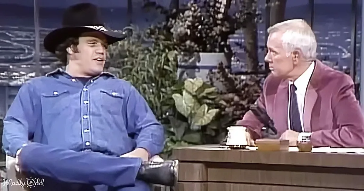 John Wayne in a cowboy hat and blue shirt sits on a couch, talking to a man in a suit on a talk show set.