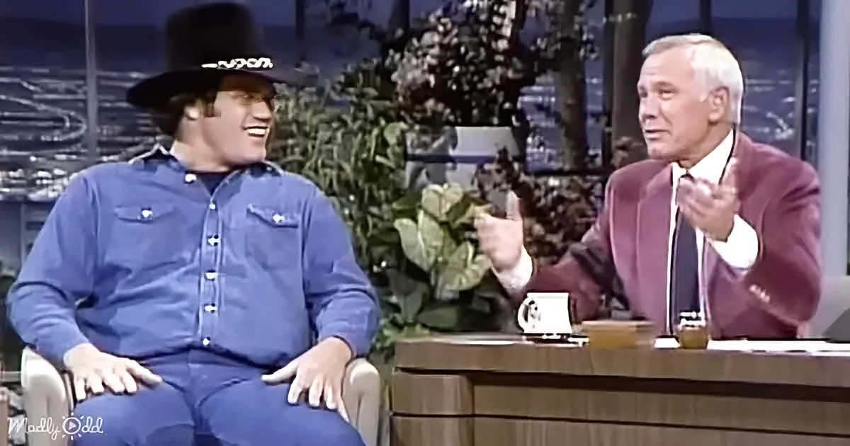 A man in a cowboy hat laughs while seated next to a man in a suit on a talk show set.