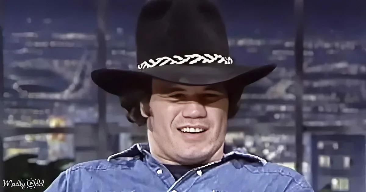 A smiling man in a blue shirt and black cowboy hat with a white braided band is featured against a cityscape backdrop.