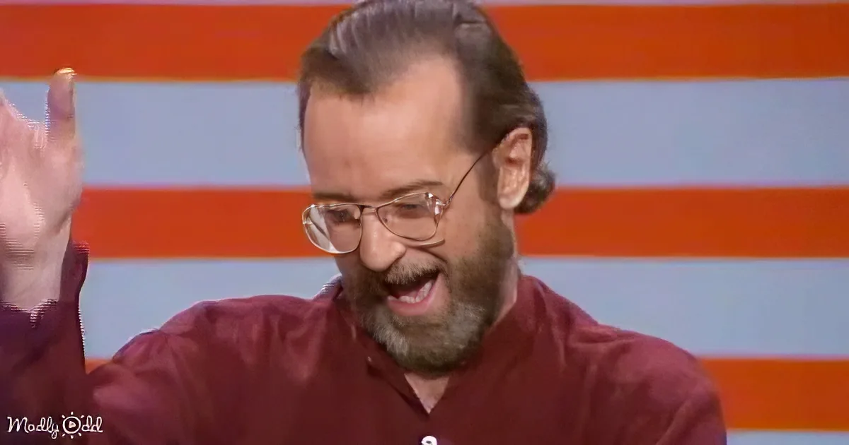 A man with a beard and glasses, wearing a maroon shirt, is smiling and raising his hand in a celebratory gesture.