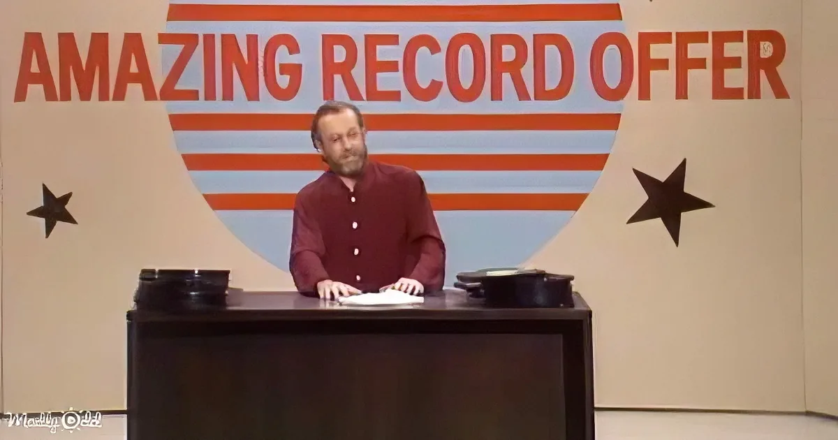 A man in a red shirt stands behind a desk with turntables, in front of a backdrop reading "AMAZING RECORD OFFER."
