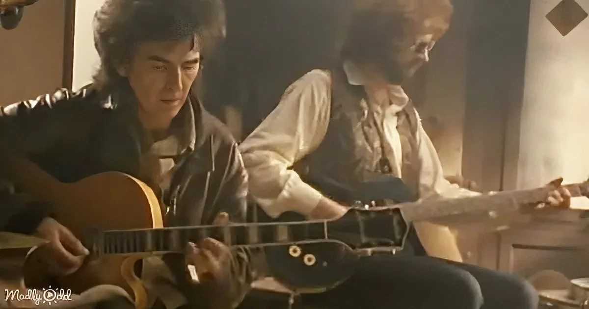 A man in a leather jacket plays a guitar, another man in period clothing sits behind him.
