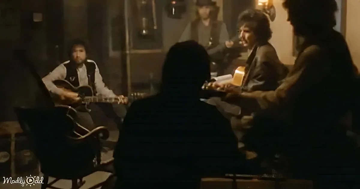 A man in a striped shirt sits facing a group of musicians playing guitars in a dimly lit room.