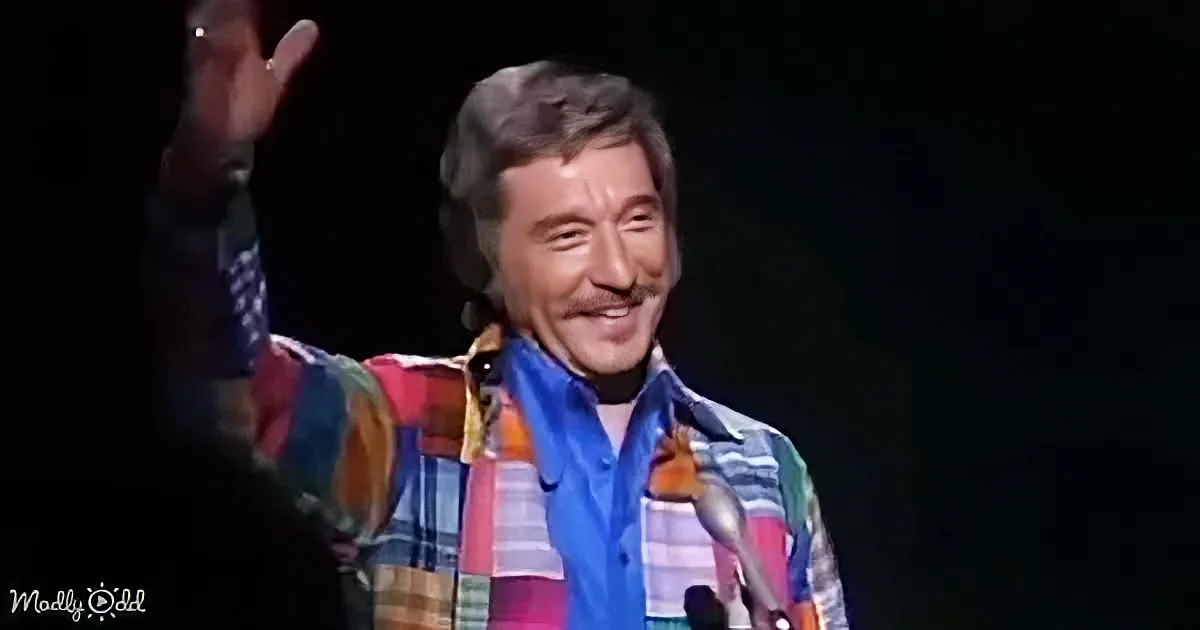 A man in a colorful plaid shirt and scarf waves while smiling on stage.